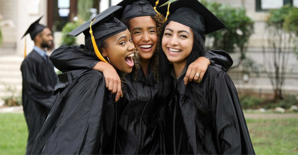 Three college graduates, two Black women and one Asian woman wearing their graduation gowns hugging and smiling.