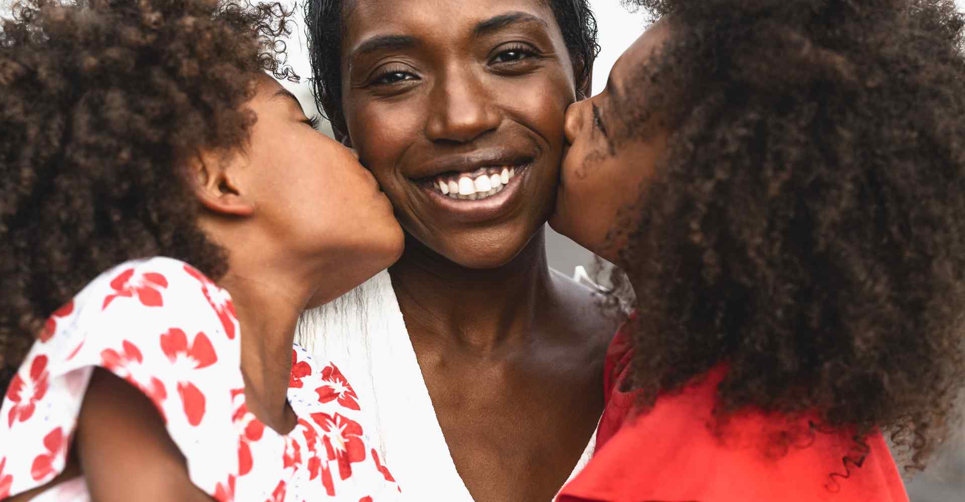 Two Black daughters kissing their moms smiling face on either side of her