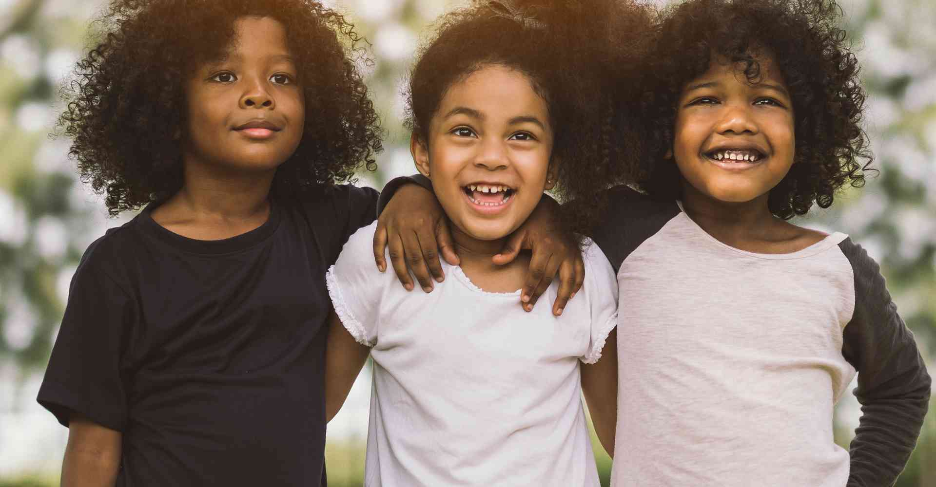 Three joyful Black kids with kinky curly hair with their arms embracing one another.