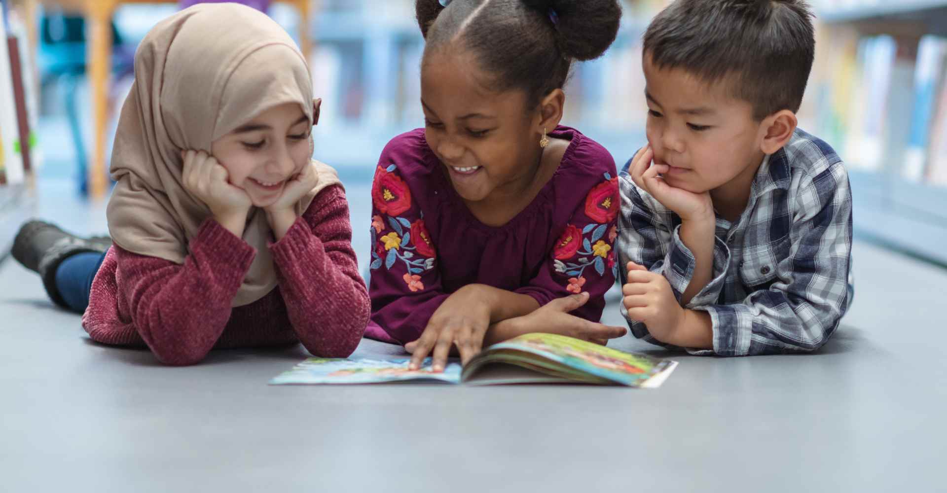 Muslim, Black, and Asian children laying on the library floor reading a book with enthusiasm.