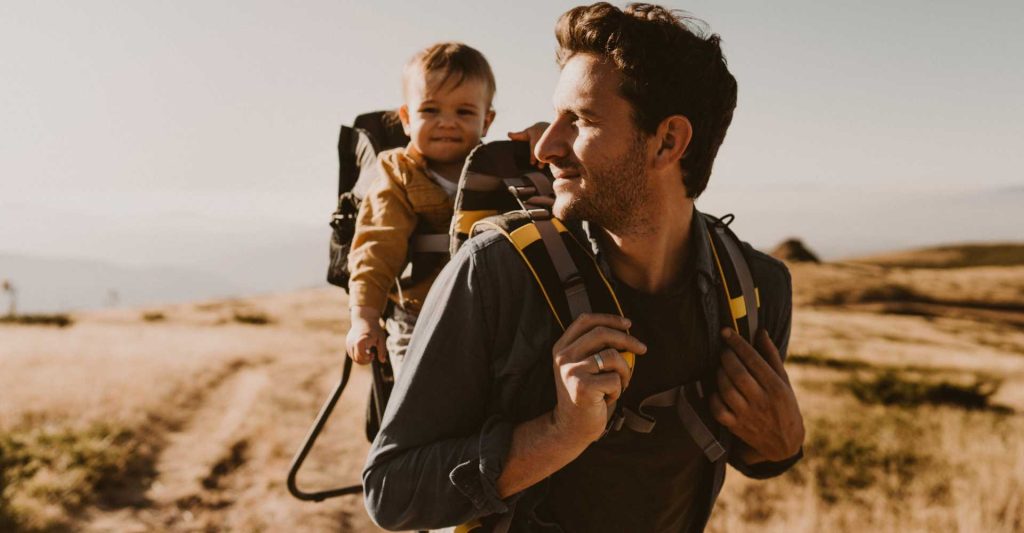 White dad looking at baby in the baby carrier on his back while the baby smiles at the camera.