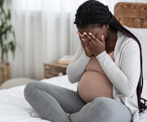 Black pregnant mom sitting on bed experiencing maternal depression with her face buried in her hands. 