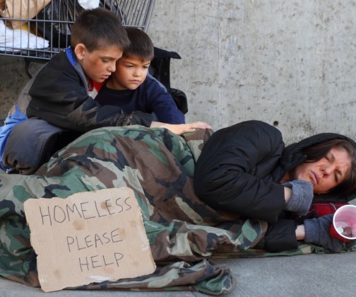 Hispanic mom laying on cement ground while her two sons comfort her experiencing homelessness, poverty, racism and toxic stress as a family. Mom is sick and sleeping while holding a near empty red solo donation cup. A sign that says "Homeless Please Help" is resting on the moms legs while she sleeps. 