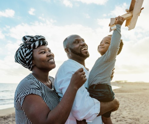 Black parents holding their baby son on the beach smiling while the son holds a wooden airplane in the air.