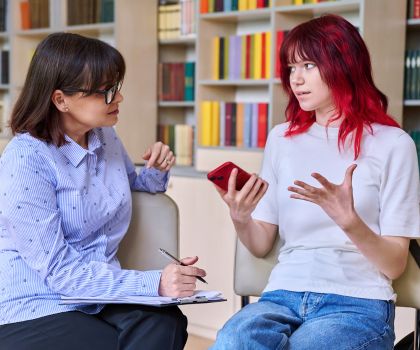 White Highschool student discussing community behavioral health needs as they relate to her with her white school counselor while holding her phone in hand. 