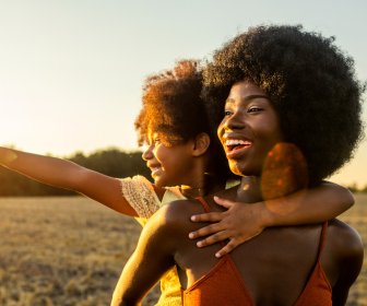 Joyful Black daughter on moms back in the middle of a sunflower field during sunset both laughing while the daughter points using her arm.