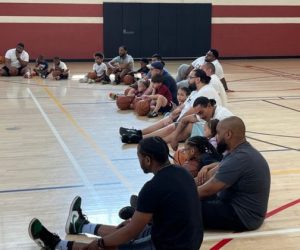 A group of diverse fathers sitting with their children, holding basketballs on the floor of a gymnasium at IFCI's 2023 Daddy & Me Day Event.