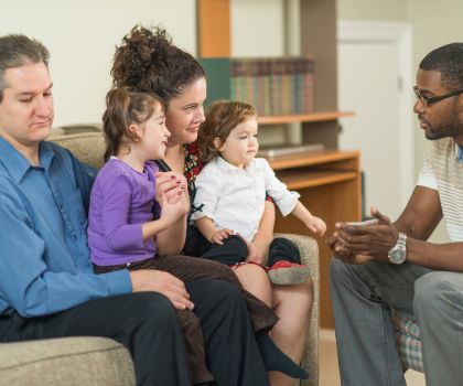 Black therapist leading therapy session on reducing ACEs with white parents and two kids sitting on couch across from him.