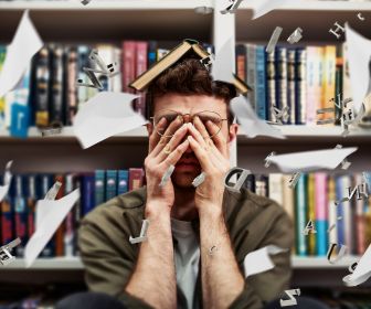 Overwhelmed college student with chaotic papers and letters flying around his head. He is rubbing his eyes with his hands because of negative student mental health.