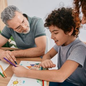 A white therapist sitting next to a little boy drawing.