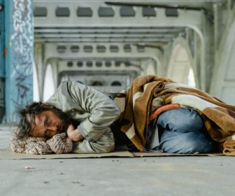 A white man experiencing homelessness laying on the cement under a bridge.