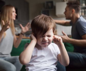 Young white boy sitting in front of his arguing parents plugging his ears with a distressed face.